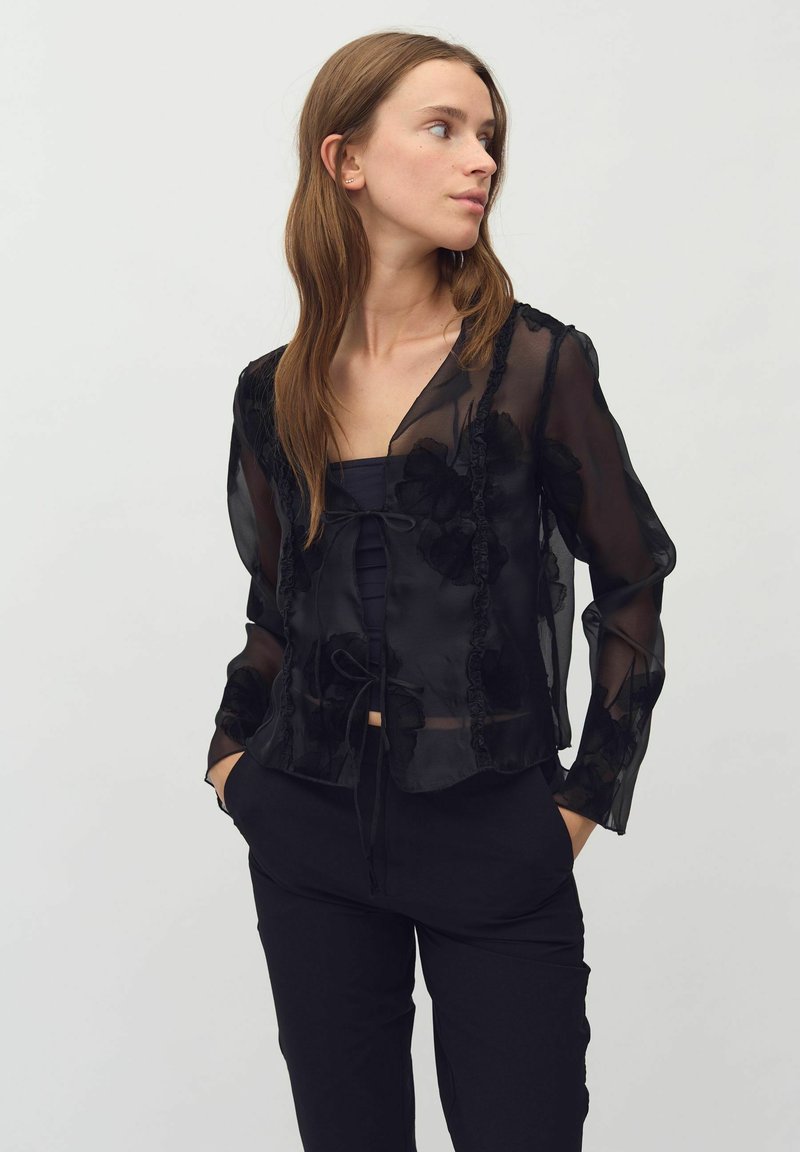 Bluză neagră transparentă cu model floral, margini volănașe și detaliu cu siret. Mâneci lungi, material ușor, suprapusă peste un top închis la culoare.