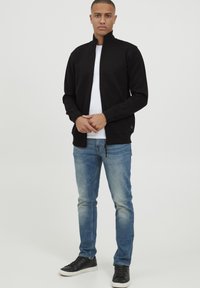 Čierna bunda na zips s vysokým golierom, spárovaná s bielym tričkom a svetlomodrými džínami. Čierne tenisky dopĺňajú outfit.