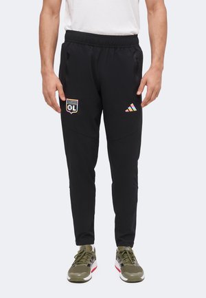 Uomo che indossa pantaloni sportivi neri con i loghi dell'Olympique Lyonnais e i loghi multicolore di Adidas, abbinati a scarpe da ginnastica verde oliva e una camicia bianca.