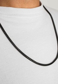 Cercea din lanț de metal negru, cu un design răsucit, așezată pe un tricou alb cu guler rotund, evidențiind textura sa netedă și forma elegantă.