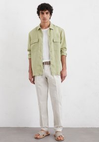 Chemise vert clair à boutons avec deux poches poitrine, t-shirt blanc à col rond, pantalon beige et sandales beiges avec un détail en nœud.