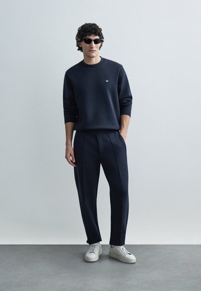 Tmavomodrá mikina s okrúhlym výstrihom a jemným logom, v kombinácii s rovnakými plisovanými nohavicami. Bielymi teniskami je outfit doplnený.