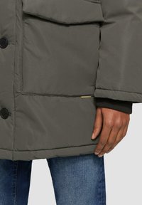 Grijze pufferjack met een boxy design, voorzien van een frontsluiting met knopen, twee voorzakken en een kleine logo-accent op de zoom.