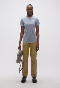 Γυναίκα όρθια, φοράει γκρι μπλουζάκι, χακί παντελόνι με δυνατότητα μετατροπής, μαύρα παπούτσια πεζοπορίας και κρατάει γκρι σακίδιο στην αριστερή της χείρα.