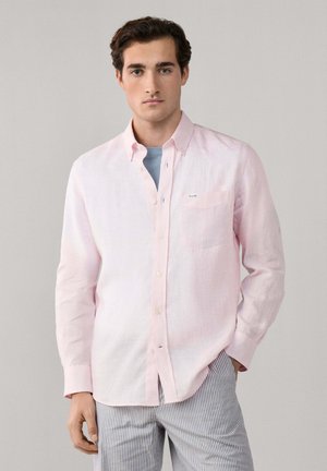Jeune homme portant une chemise à boutons rose clair sur un t-shirt bleu et un pantalon à rayures, debout devant un fond gris uni.