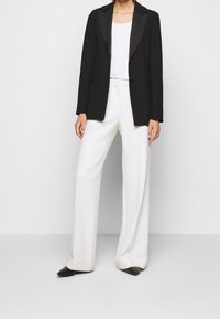 Blazer noir avec revers en satin, associé à un haut blanc et un pantalon blanc à jambe large. Des chaussures noires à bout pointu complètent la tenue.