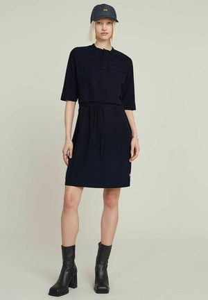 Robe bleu marine foncé avec des manches courtes, un col boutonné, une taille ceinturée, des poches latérales et un petit logo près de l'ourlet. Portée avec des bottes noires.