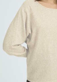 Beige stickad tröja med vid halsringning, långa sänkta ärmar och en mjuk, texturerad finish. Avslappnad passform som är lämplig för lager-på-lager.