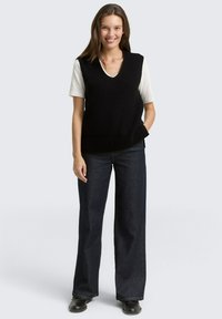 Schwarze Strickweste mit V-Ausschnitt, über einem weißen Hemd getragen. Das Modell trägt dunkle Jeans mit weiten Beinen und schwarze Schuhe.