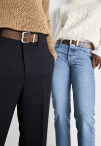 Brun lærreim med sølvspenne, båret over marineblå plisserte bukser og lyseblå jeans, kombinert med strikkede beige og kremfargede gensere.