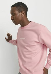 Hele roosa sweatshird, millel on soonikkoesed ja kaelus, ning millel on valge tekst "Originals Studio". Sile kangas ja mugav lõige.