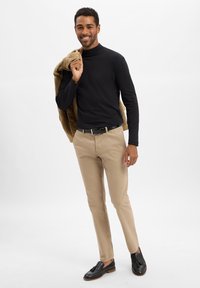 Czarny golf, beżowe spodnie i czarne mokasyny z frędzlami. Model trzyma na ramieniu jasnobrązową pikowaną kurtkę.