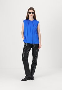 Top senza maniche blu con pieghe, abbinato a pantaloni aderenti neri e lucidi e stivali con tacco neri; la modella indossa occhiali da sole neri.