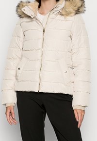 Kvinna iklädd en krämfärgad puffjacka med huva i päls, framzipper och knäppningsfickor, kombinerad med svarta byxor.