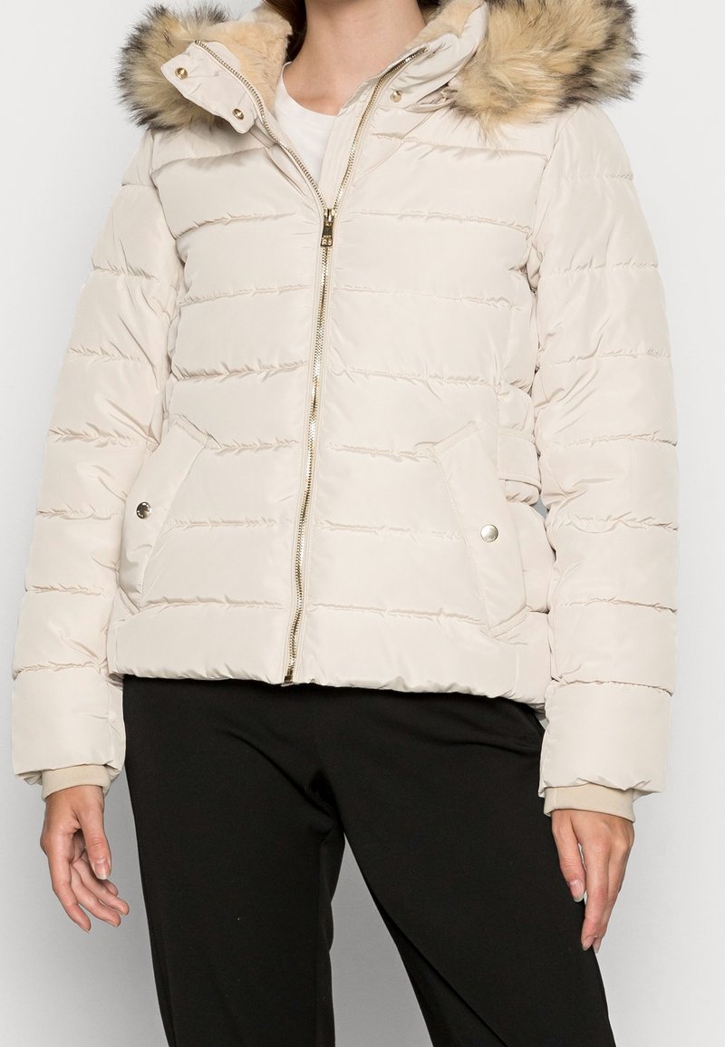 Kvinna iklädd en krämfärgad puffjacka med huva i päls, framzipper och knäppningsfickor, kombinerad med svarta byxor.