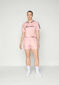 Conjunto atlético rosa feito de um material suave, apresentando uma t-shirt recortada com logotipo preto e detalhes em baixa, emparelhada com calções a condizer. Ténis brancos.