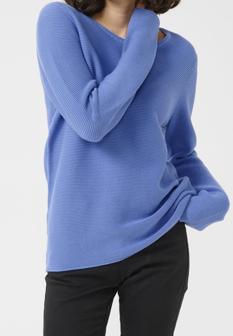 Suéter de punto azul con canalé, cuello en V y mangas largas, con un corte relajado y textura sutil, acompañado de pantalones negros.