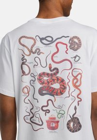 Camiseta blanca con diversas ilustraciones de serpientes coloridas y etiquetas detalladas, incluida un personaje de oso con un sombrero en la parte inferior.