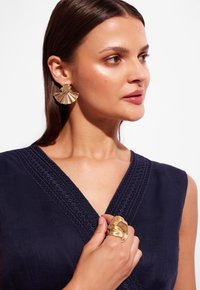 Złote kolczyki w kształcie wachlarza, teksturowana granatowa sukienka z ozdobnym dekoltem oraz oversize'owy złoty pierścionek. Gładka skóra i neutralny makijaż.