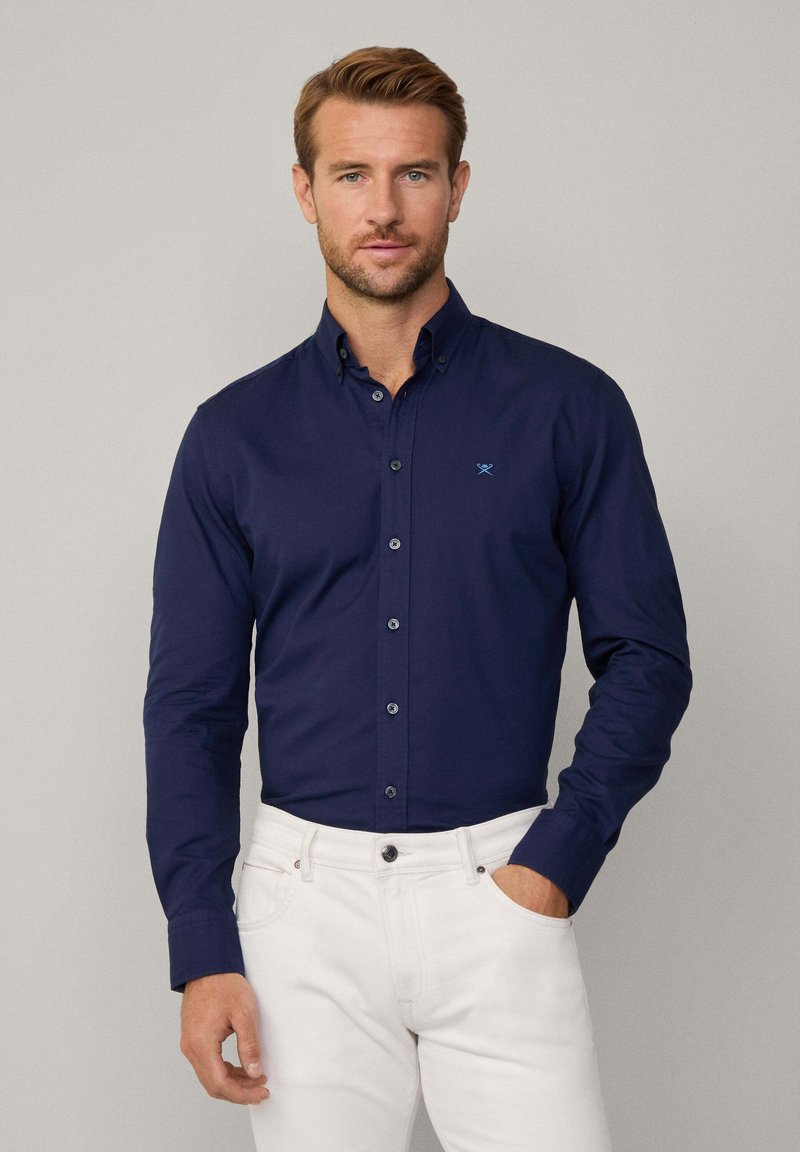 Marineblauwe overhemd met knoopjes en een kraag, voorzien van een klein blauw logo op de linkerborst. Gestoeld met witte broeken. Gladde textuur.
