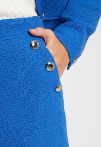 Tessuto blu texturizzato con un motivo sottile, caratterizzato da bottoni metallici ai lati e una tasca, che accentuano un design strutturato.