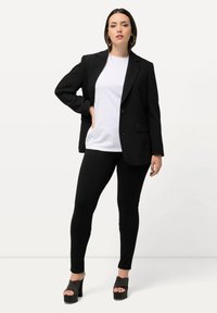 Zwarte blazer over een wit T-shirt, gecombineerd met zwarte leggings en hoge hakken. De blazer heeft een enkele knoop en twee zijzakken.