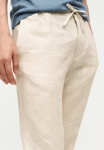 Άνδρας που φοράει μπεζ παντελόνι από λινό με κορδόνι και μπλε πουκάμισο, εμφανίζοντας τον κορμό και το πάνω μέρος των ποδιών του με φόντο μια απλή επιφάνεια.
