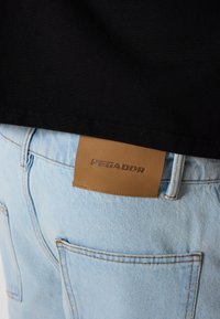 Person trägt hellblaue Jeans mit braunem Pegador-Label und ein schwarzes Shirt, das teilweise den Bund und die Gesäßtaschen bedeckt.