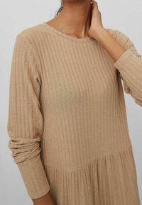 Robe beige côtelée à manches longues, avec un col rond et une jupe froncée. Tissu doux avec une finition texturée.