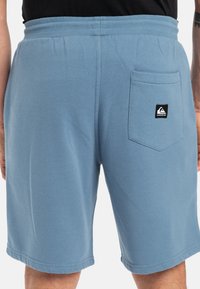 Lichtblauwe shorts van zachte stof, met een elastische tailleband en een achterkantzak met een zwart merklogo. Glad textuur, eenvoudig ontwerp.