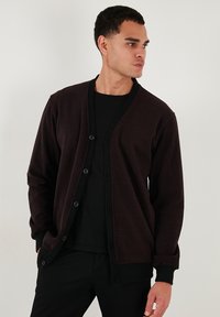 Cardigan marrone scuro realizzato in soffice tessuto, con scollatura a V, chiusura a bottoni, maniche lunghe e rifiniture nere lungo i bordi.
