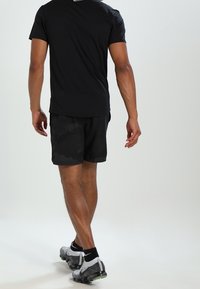 Camiseta deportiva negra de diseño ajustado, combinada con pantalones cortos negros con estampado. El conjunto se completa con zapatillas deportivas en gris y negro.