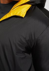 Svart vadderad jacka med en glänsande yta, med en kontrasterande gul huva och dragkedjedetalj. Fokusera på axel- och krageområde.