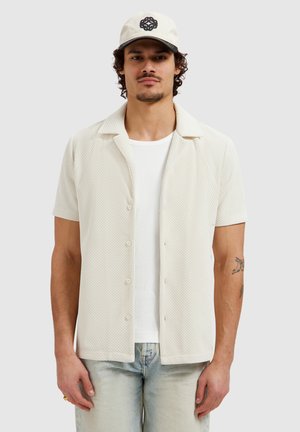 Man wearing cream textured short-sleeve button-up shirt over white tee, light wash jeans, cream cap with black logo, and visible tattoos on arms.