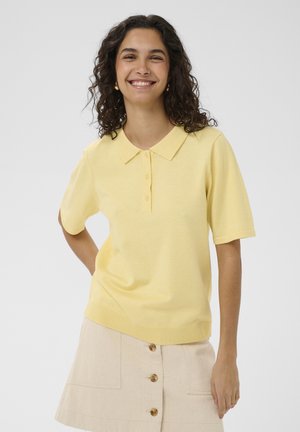 Usmievavá žena s kučeravými vlasmi, oblečená v svetložltom polo tričku s krátkym rukávom a krémovej sukni so zapínaním na gombíky, stojaca pred jednoduchým pozadím.