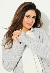 Světle šedý žebrovaný cardigan s uvolněným střihem, s dlouhými rukávy a texturovaným vzorem. Nosený přes bílou košili na knoflíky.