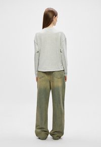 Mulher com cabelo castanho comprido, usando brincos de argola prateados, uma blusa de mangas longas cinza claro e calças largas verdes desbotadas, de costas.