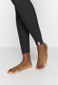 Leggings neri realizzati in materiale elasticizzato, con un design aderente, lunghi fino alla caviglia e un piccolo tag con il marchio vicino all'orlo.