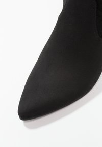 Czarny zamszowy botek na kostkę z ostrym noskiem i gładką fakturą. Charakteryzuje się eleganckim, minimalistycznym designem bez widocznych okuć czy dodatków.