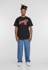 Melnas īsām piedurknēm T-krekls ar sarkanu un baltu uzrakstu "Rocawear 99". Apvienots ar gaiši ziliem platajiem džinsiem un baltiem sporta apaviem.