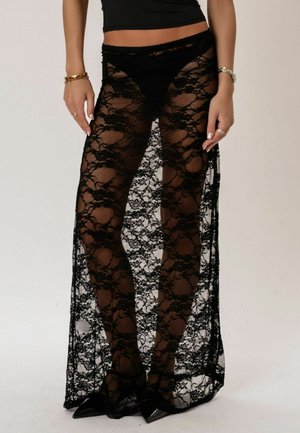 Frau trägt durchsichtige, schwarze Spitzenhose mit weitem Bein über schwarzer Unterwäsche und spitzen, schwarzen Absätzen vor neutralem Hintergrund.