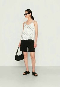 Top branco sem mangas com pequeno padrão floral colorido, combinado com calções pretos. A modelo usa óculos de sol pretos e traz uma bolsa preta.