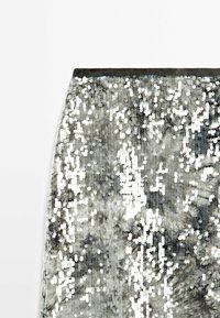 Tissu en sequins argentés avec une texture réfléchissante, présentant un design scintillant sur un fond sombre contrasté. Les bords sont droits et nets.