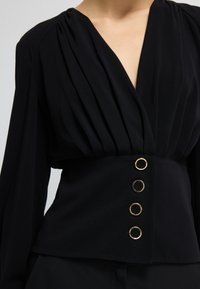 Blusa nera con design plissettato, scollatura a V profonda, maniche lunghe e bottoni circolari dorati lungo la vita. Tessuto dalla consistenza liscia.