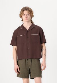 Braunes Kurzarmhemd mit weißen Paspeln, zwei Fronttaschen und schwarzen Knöpfen, kombiniert mit olivgrünen Shorts.