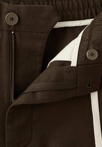 Gros plan d’un pantalon marron foncé avec un bouton, une braguette à zip et une doublure intérieure blanche visible à travers la fermeture éclair ouverte.