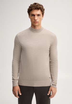 Junger Mann mit kurzem lockigem Haar, der einen hellbeigen Pullover mit langen Ärmeln und dunkle Hose trägt, steht vor einem einfarbigen hellen Hintergrund.