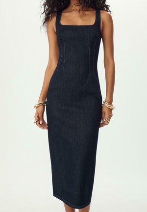 Ærmeløs mørk denim midi-kjole med firkantet halsudskæring, figursyet talje, båret med gyldne armbånd og ringe på begge hænder.