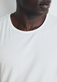 White cotton t-shirt with a wide round neckline, smooth texture, and visible stitching around the collar. Close-up of the upper body.