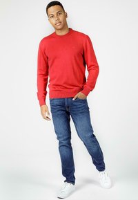 Czerwony sweter z długim rękawem i okrągłym dekoltem, w połączeniu z ciemnoniebieskimi dżinsami i białymi trampkami, prezentujący swobodny, dopasowany styl.